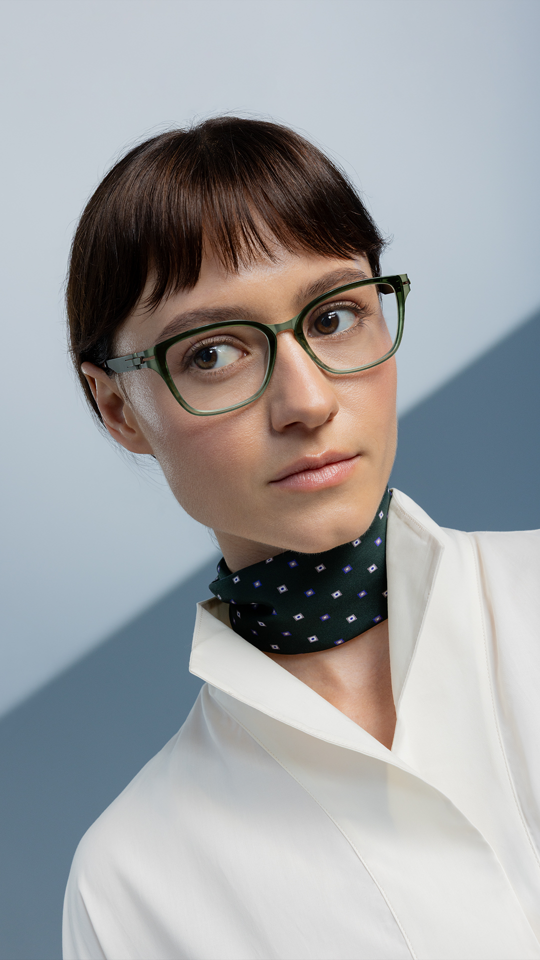 Woman in a white blouse and patterned green scarf wearing OVVO Optics DUO Marla translucent green eyeglasses against a blue-gray background