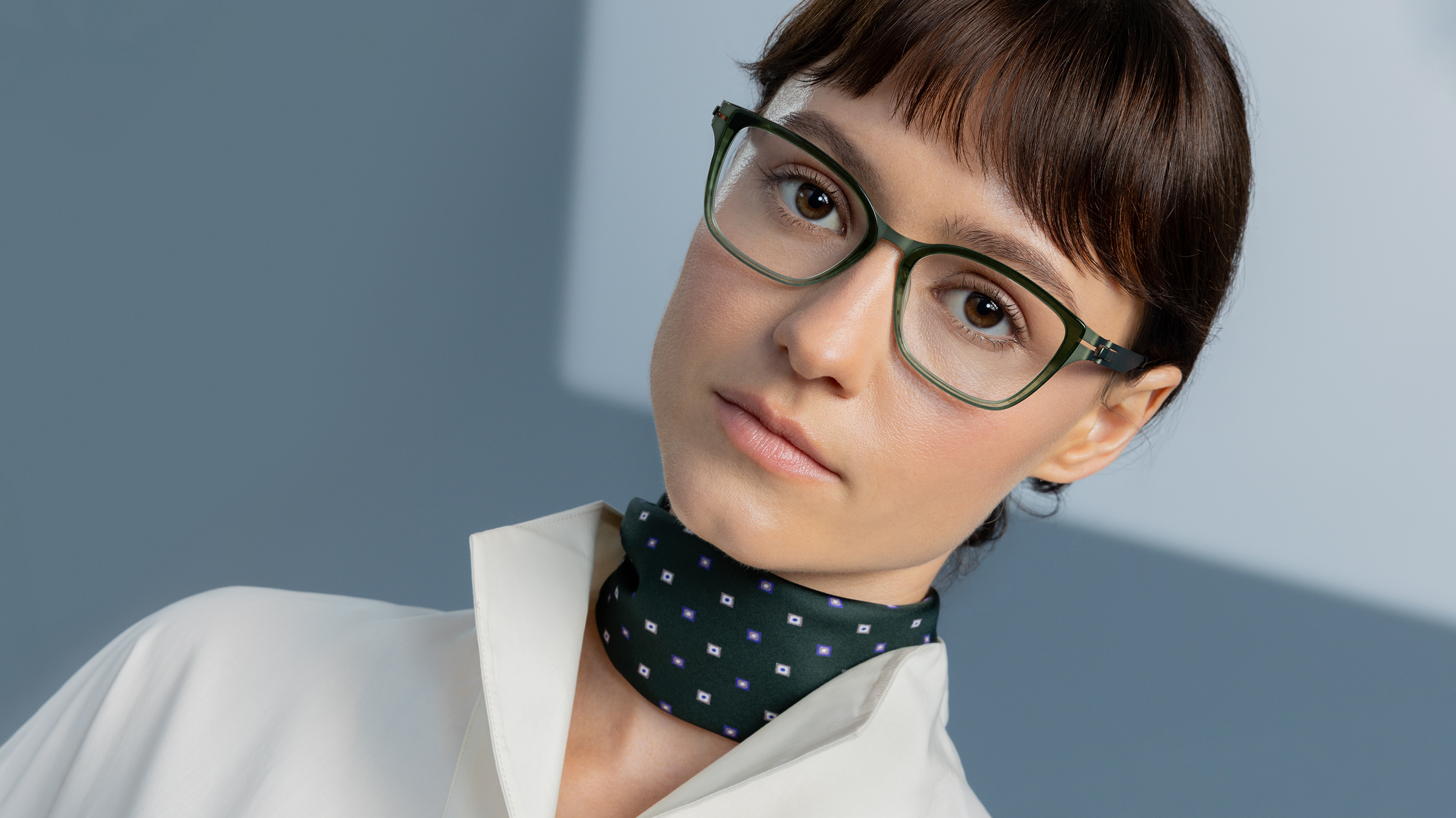 Woman in a white blouse and patterned green scarf wearing OVVO Optics DUO Marla translucent green eyeglasses against a blue-gray background